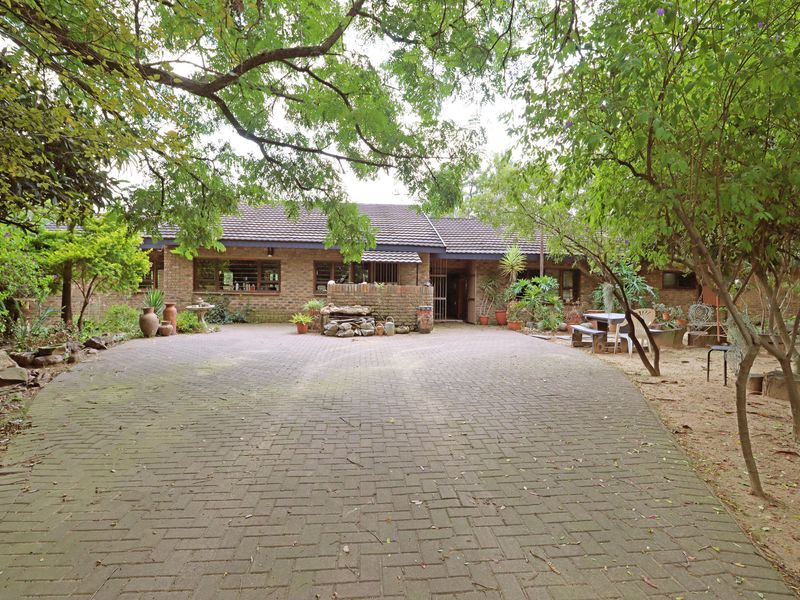 Front view of a single-story brick farmhouse featuring a paved driveway, mature trees, and a tiled roof, set within a landscaped garden area.