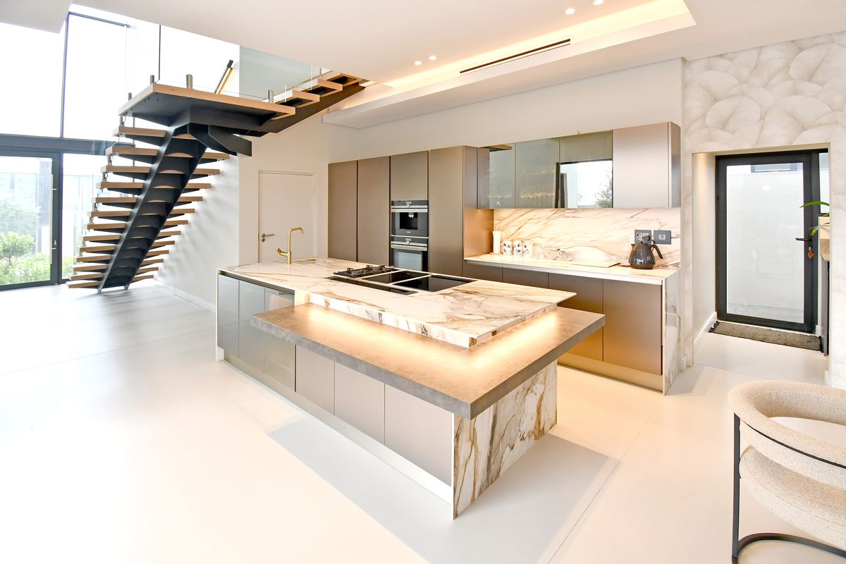 Modern open-plan kitchen featuring a marble island with integrated hob, high-end built-in appliances, and a striking floating staircase with glass balustrade.