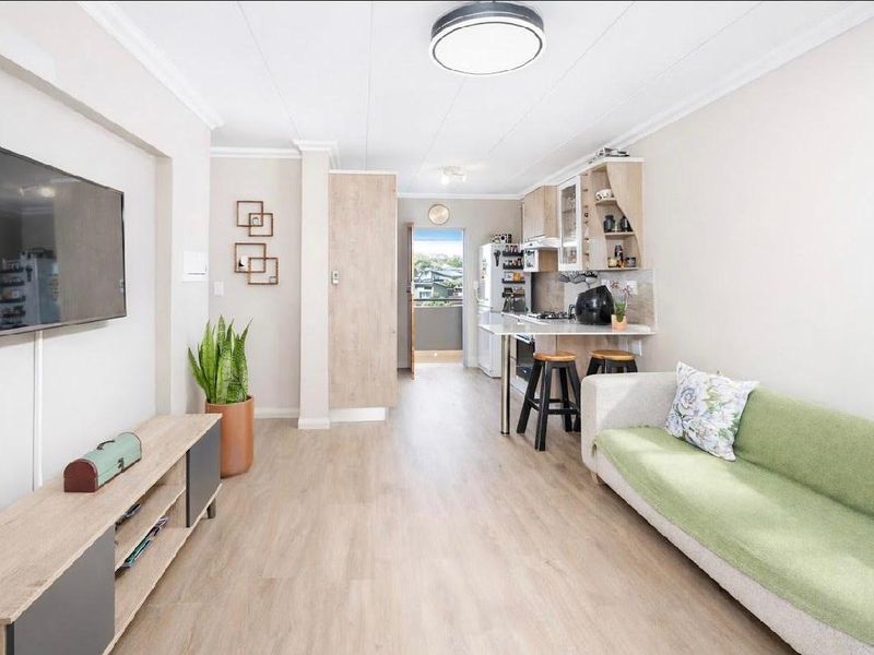An open-plan living and kitchen area featuring light wood-look laminate flooring, modern cabinetry, and a breakfast bar, leading to an outdoor balcony.