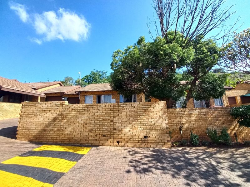 Front exterior view of a face-brick townhouse featuring a paved driveway and a prominent boundary wall with mature trees.