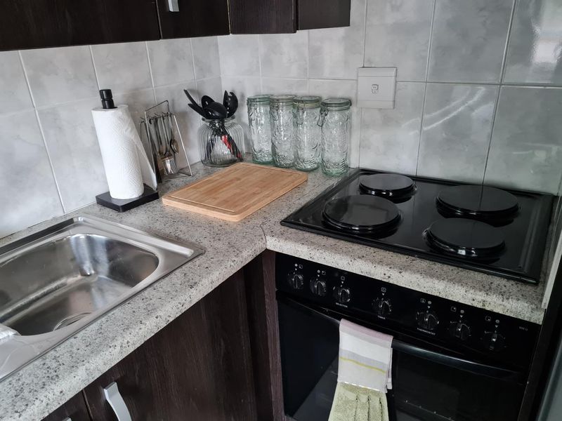 A compact kitchen area featuring dark wood cabinetry, a granite-look laminate countertop, a stainless steel sink, and a built-in electric oven with hob.