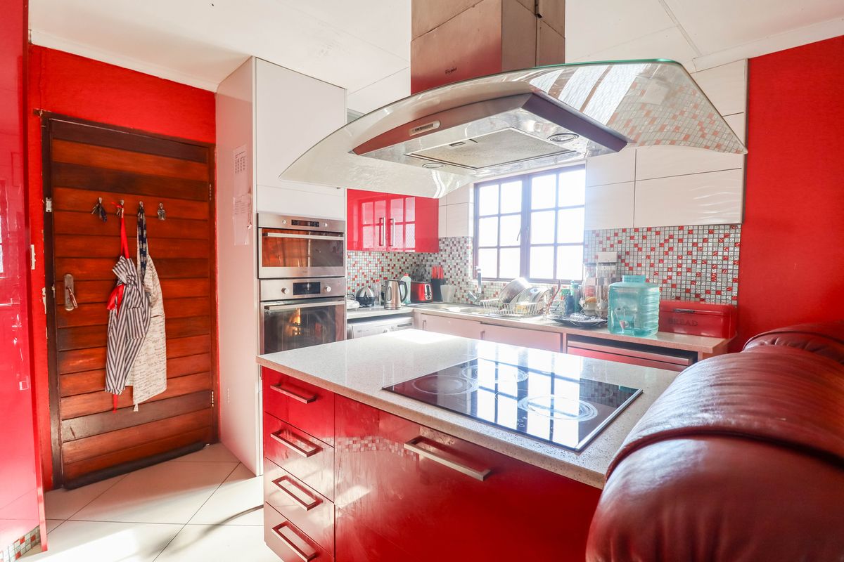 A vibrant kitchen featuring a central island with a built-in cooktop, modern extractor fan, built-in oven, and striking red cabinetry with mosaic tile accents.
