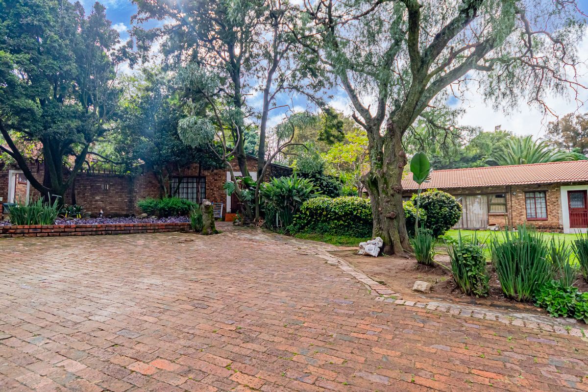 A spacious rear exterior view of the cottage featuring a brick-paved courtyard, mature trees, and established garden beds against a classic facebrick facade.