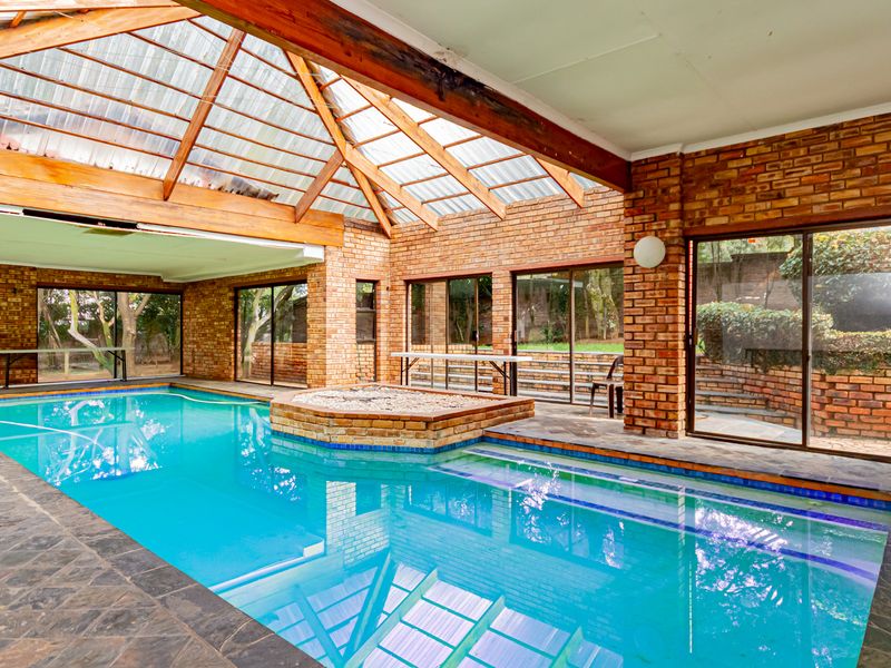 Spacious indoor swimming pool area featuring exposed brick walls, a timber-framed translucent roof, and large sliding glass doors opening to the garden.