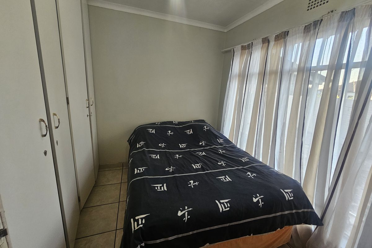 A functional bedroom featuring built-in cupboards, tiled flooring, and large windows with curtain rails, offering a clean and practical living space.