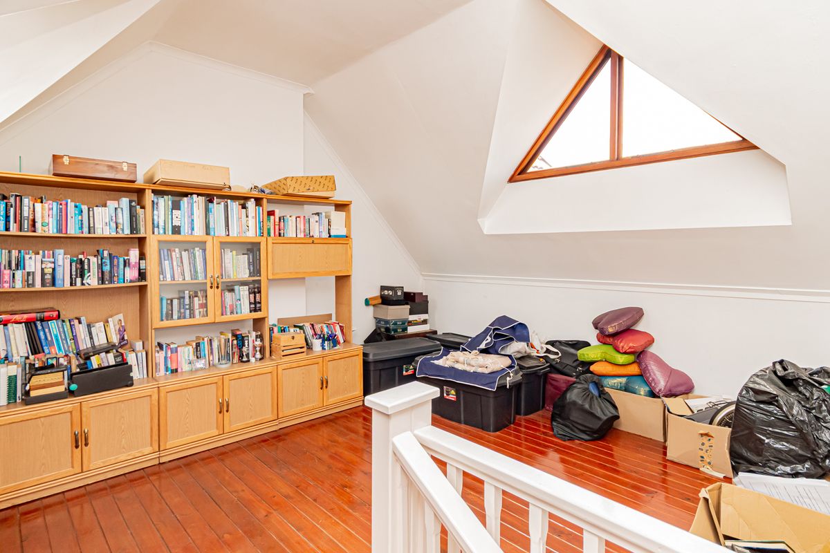 An attic-style bedroom featuring sloped ceilings, a unique triangular window, and polished timber flooring, currently utilized as a storage and study area.