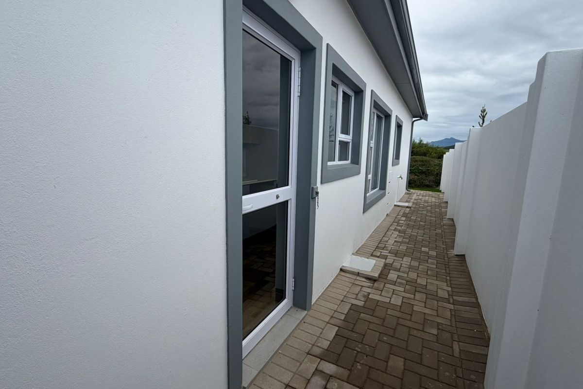 A clean side exterior view of the property featuring a paved walkway, white boundary wall, and modern aluminium window and door frames.