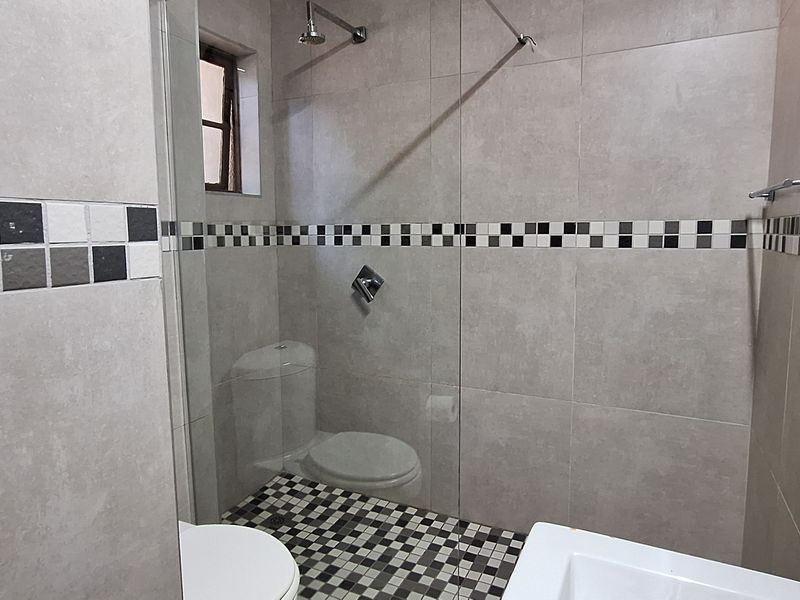 A clean, modern bathroom featuring a glass-enclosed shower, neutral wall tiling with a decorative mosaic border, and a functional toilet.
