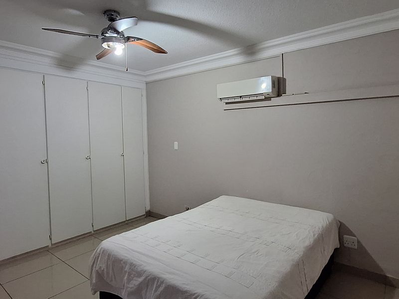 A functional bedroom featuring tiled flooring, built-in cupboards, a ceiling fan, and a wall-mounted air conditioning unit.