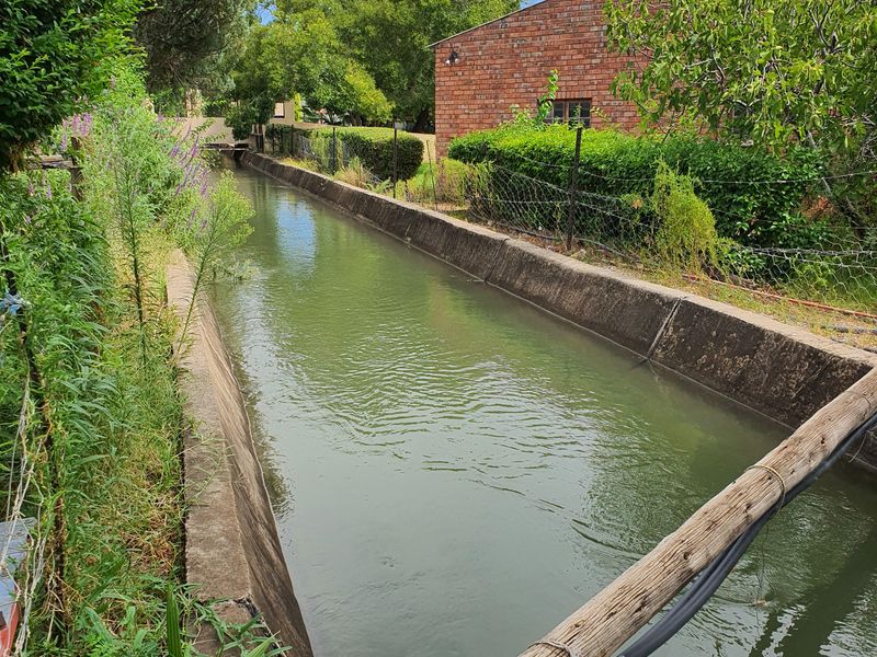 View of a concrete irrigation canal running alongside a brick boundary wall and landscaped garden area.
