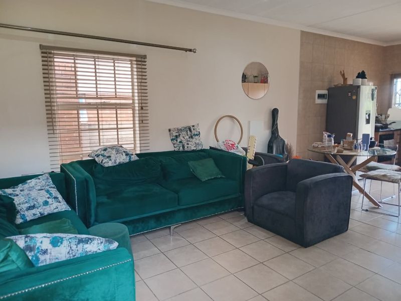 Open-plan living area featuring tiled flooring and neutral walls, seamlessly connecting to a kitchen space with tiled splashback and natural light.