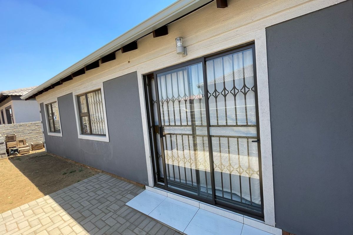 Rear exterior view of the house featuring a paved patio area, sliding glass door with security gate, and modern grey exterior wall finish.