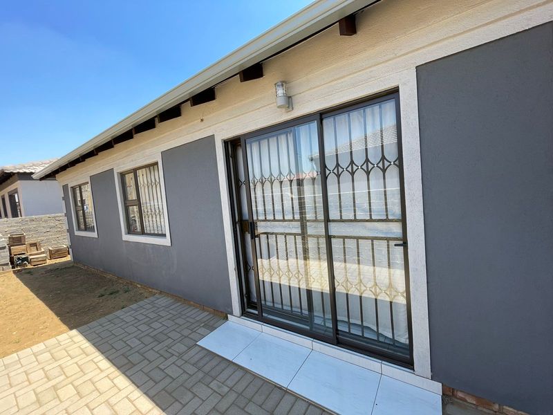 Rear exterior view of the house featuring a paved patio area, sliding glass door with security gate, and modern grey exterior wall finish.
