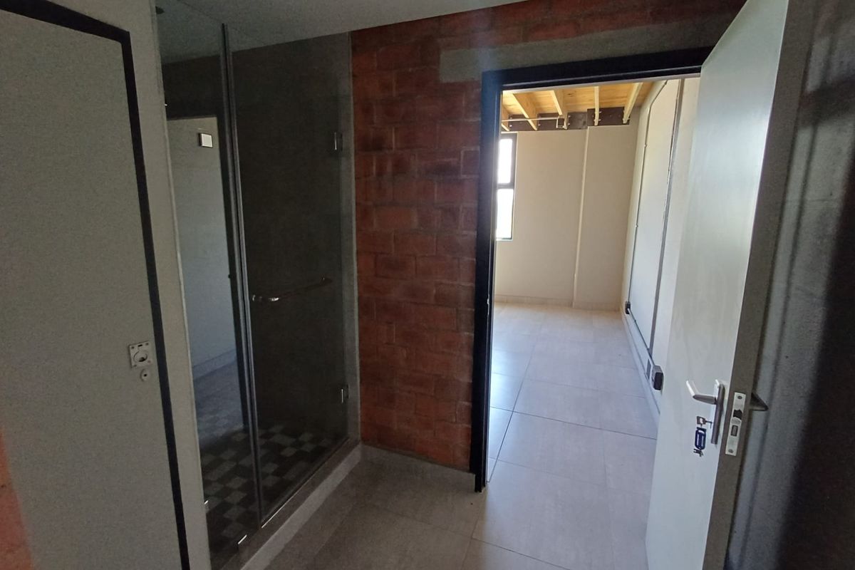 An industrial-style bathroom featuring a glass shower enclosure, exposed brickwork, and tiled flooring, leading into an adjacent unfinished living space.