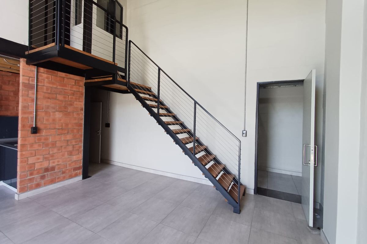 Modern open-plan living area featuring an industrial-style steel staircase, mezzanine level, exposed brick accent wall, and neutral tiled flooring.