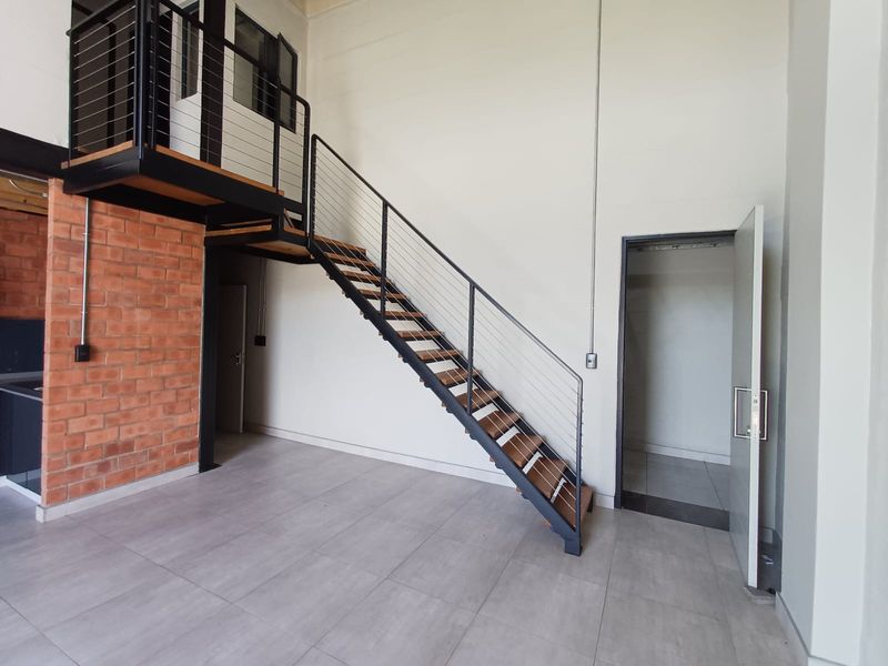Modern open-plan living area featuring an industrial-style steel staircase, mezzanine level, exposed brick accent wall, and neutral tiled flooring.