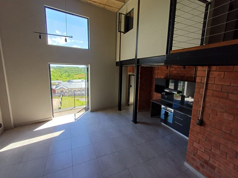Spacious open-plan living area featuring double-volume ceilings, exposed brickwork, a modern kitchen with built-in appliances, and access to a balcony with scenic views.