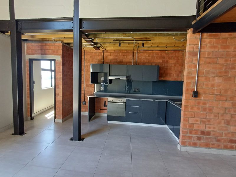 Modern industrial-style kitchen featuring exposed brickwork, steel beams, sleek dark cabinetry, and integrated oven, set within an open-plan apartment layout.