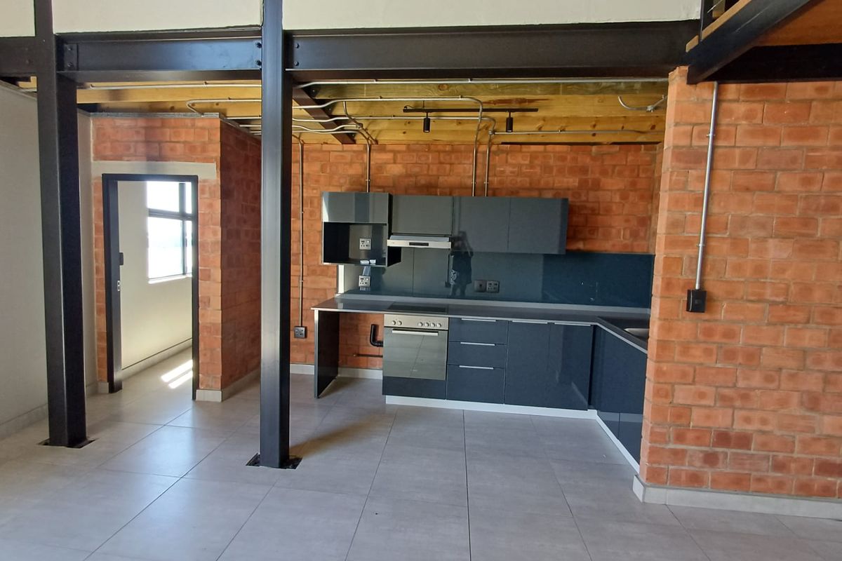 Modern industrial-style kitchen featuring exposed brickwork, steel beams, sleek dark cabinetry, and integrated oven, set within an open-plan apartment layout.