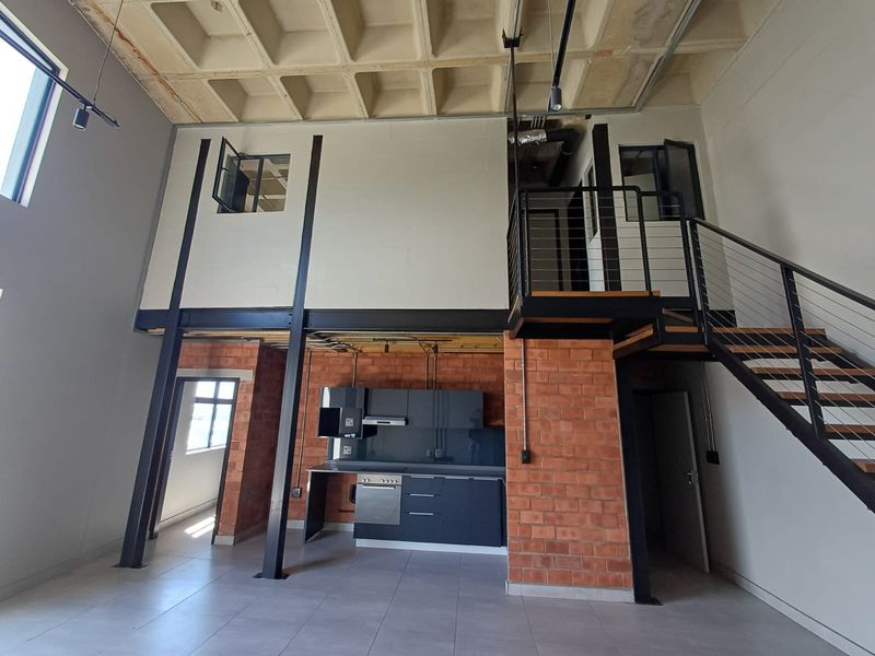 Spacious industrial-style open plan living area featuring exposed brickwork, a waffle concrete ceiling, and a modern kitchen with integrated appliances and mezzanine staircase.
