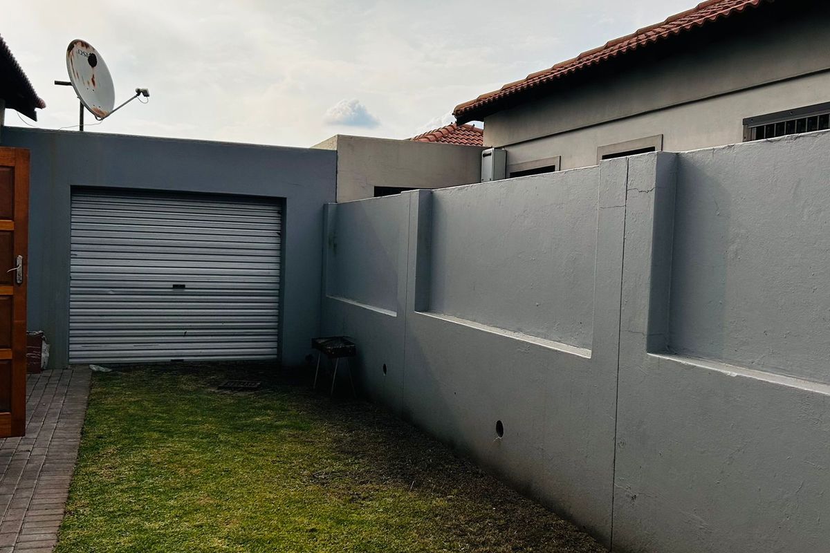 A rear exterior view of the townhouse featuring a private garden area, a roller garage door, and a perimeter boundary wall.
