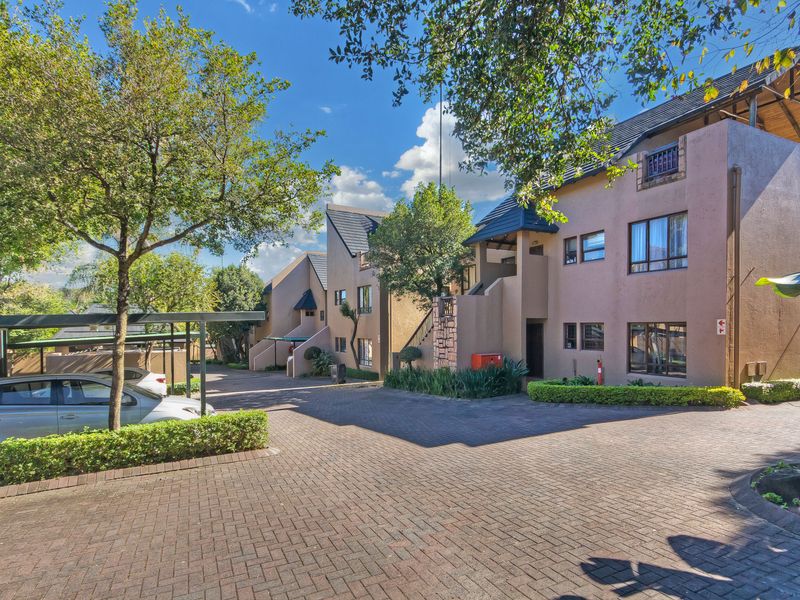 A well-maintained apartment complex exterior featuring paved driveways, covered parking bays, and established landscaped gardens under a clear blue sky.