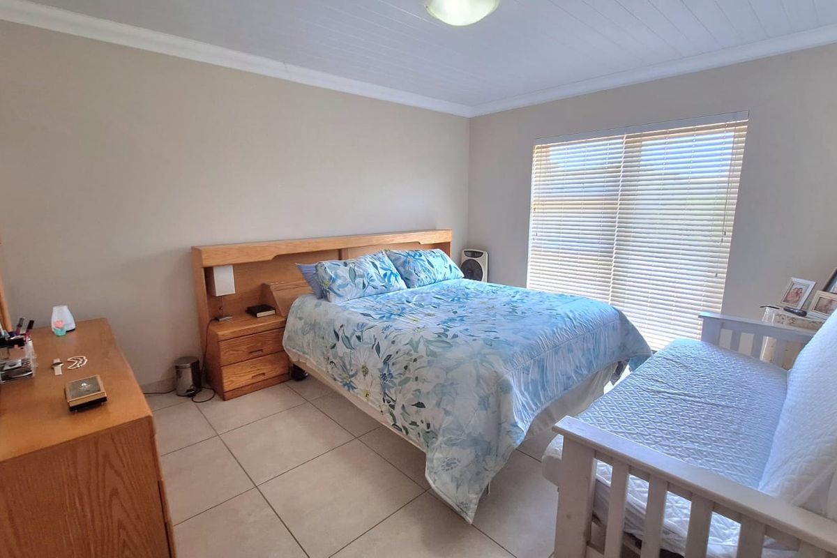 A well-lit bedroom featuring neutral tiled flooring, built-in wooden headboard with integrated bedside tables, and large window with fitted blinds.