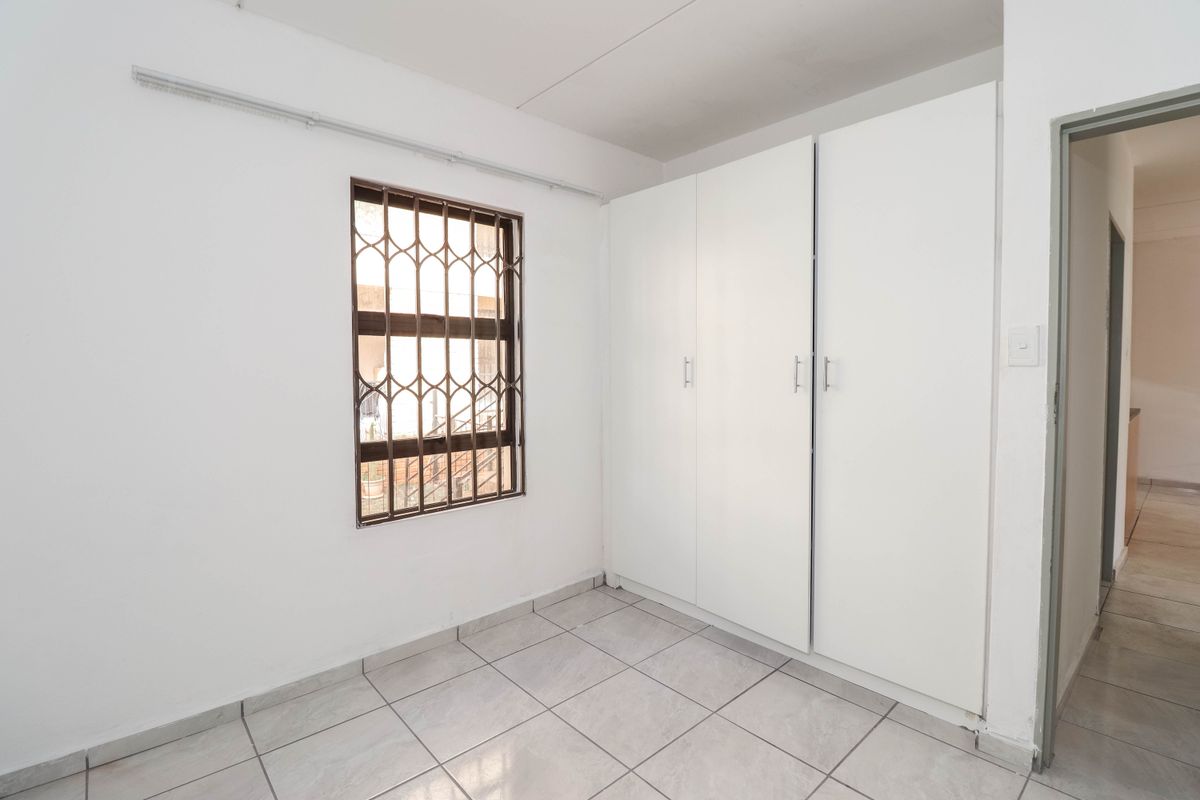 A clean, empty bedroom featuring tiled flooring, built-in cupboards, and a window fitted with security bars.