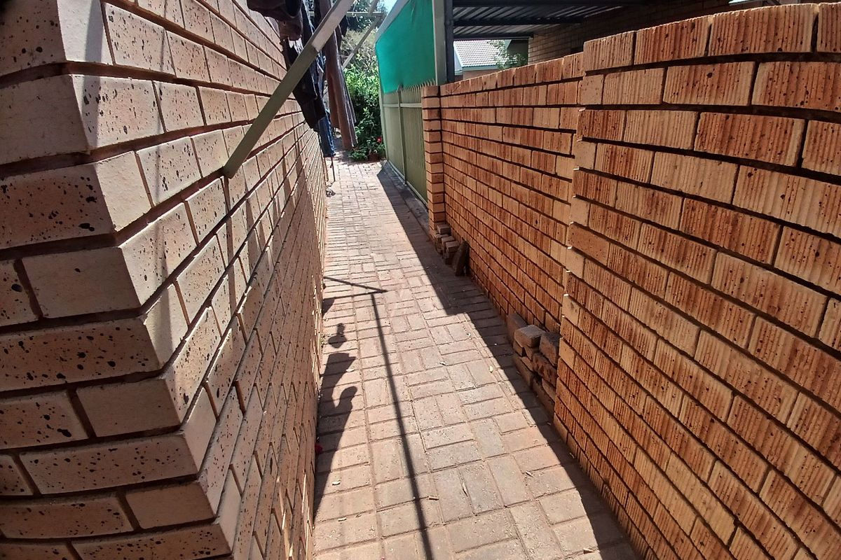 A narrow side passage featuring face brick walls and paved flooring, leading towards a rear outdoor area with a metal carport structure.