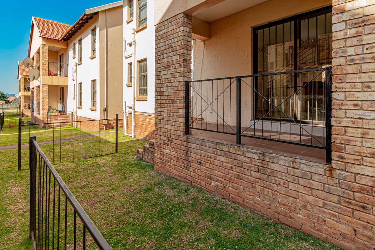 Exterior view of an apartment complex featuring a ground-floor patio with brick detailing, metal security gates, and a shared garden area.
