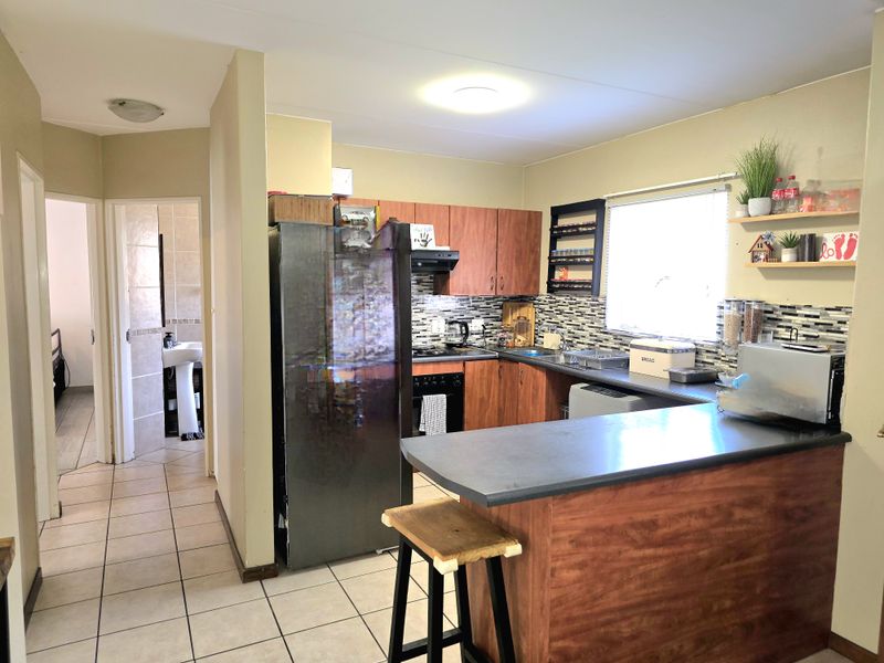 A functional kitchen featuring wood-look cabinetry, a breakfast bar, and a tiled backsplash, with an adjacent hallway leading to a bathroom.