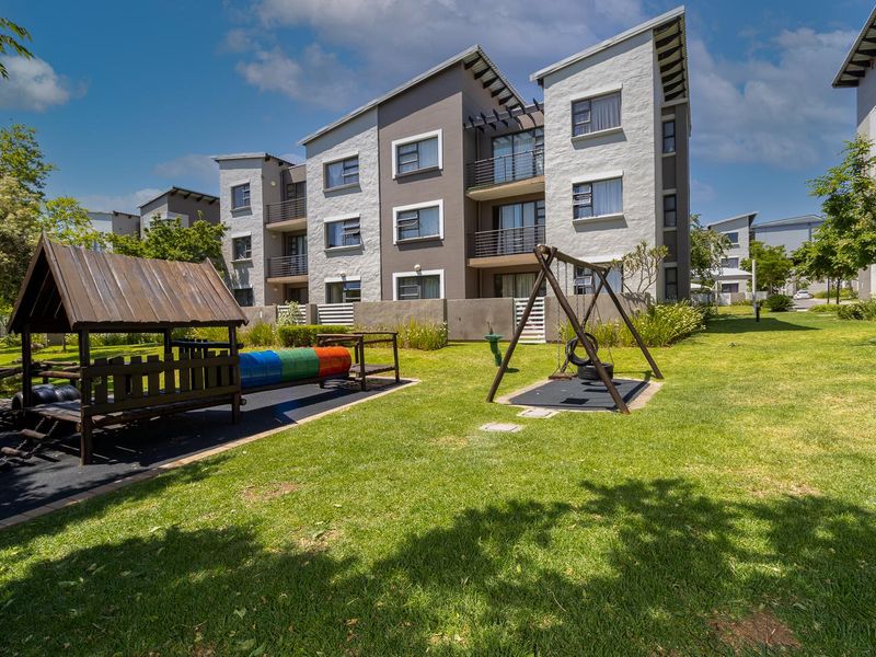 A view of the rear elevation of a modern apartment complex featuring well-maintained communal gardens and a children's play area.