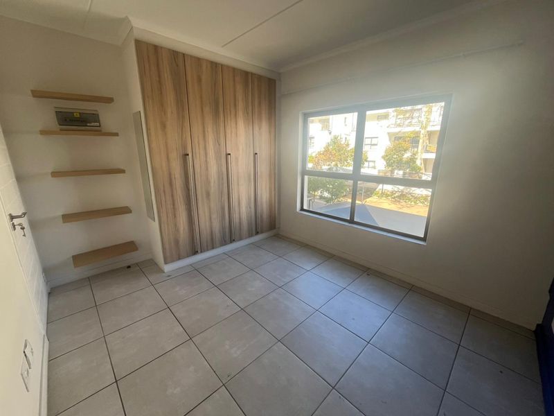 A bright, empty bedroom featuring modern wood-finish built-in cupboards, floating wall shelves, and neutral floor tiling with a large window.