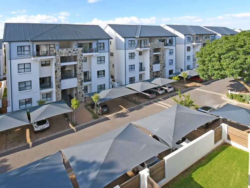 An elevated view of a modern apartment complex featuring multi-story residential buildings, paved parking areas with shade cloth carports, and landscaped grounds.