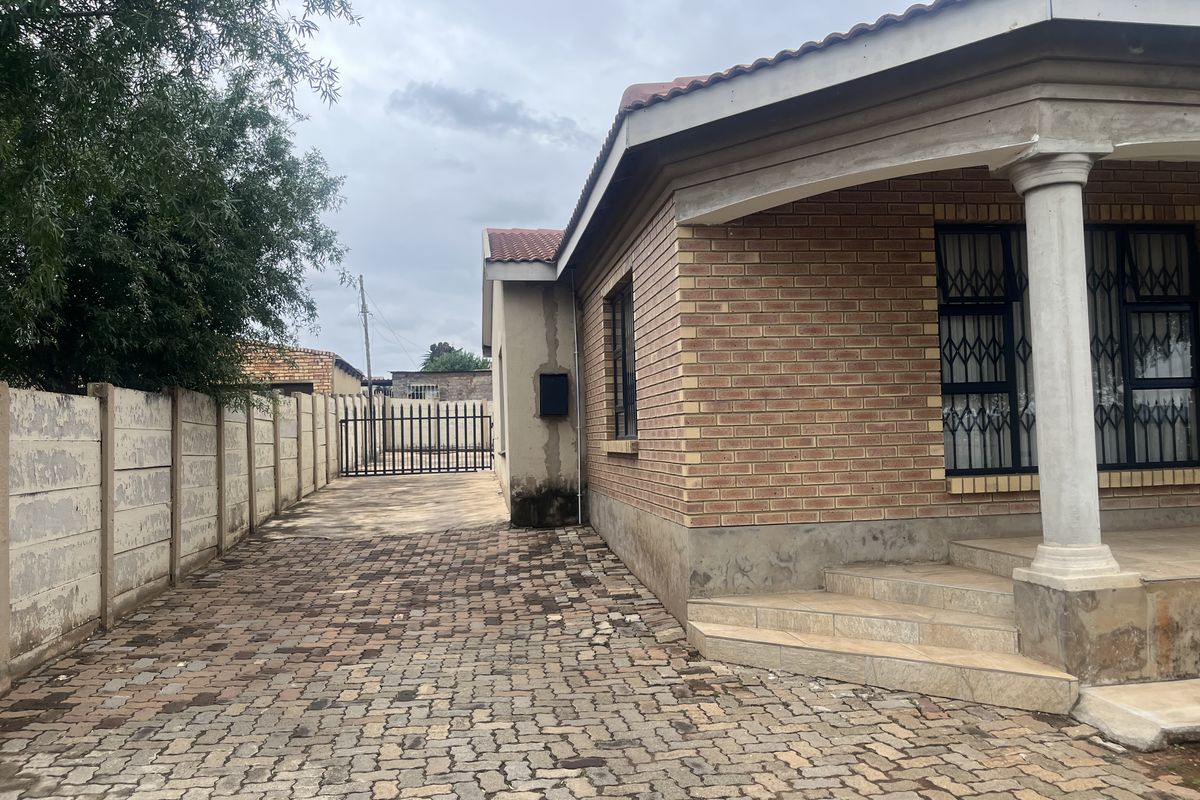Side view of a face brick house featuring a paved driveway, secure metal gate, and tiled entrance steps with concrete columns.