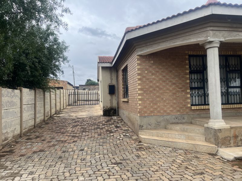 Side view of a face brick house featuring a paved driveway, secure metal gate, and tiled entrance steps with concrete columns.