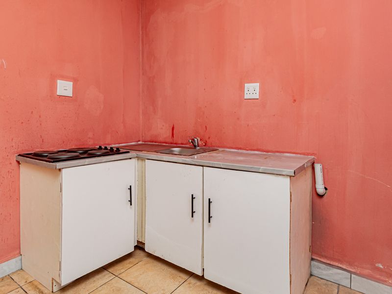 A compact kitchen area featuring basic white cabinetry, a stainless steel sink, and an electric hob, set against a plain wall with tiled flooring.