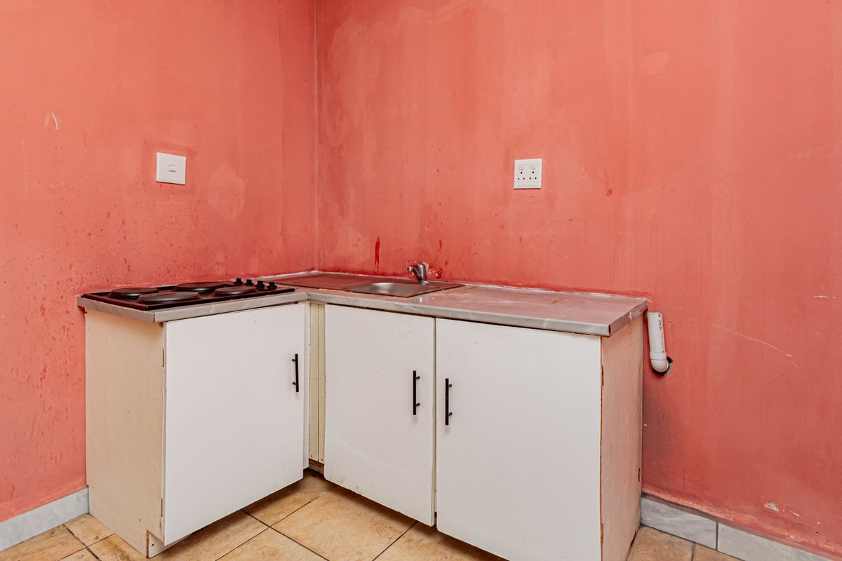 A compact kitchen area featuring basic white cabinetry, a stainless steel sink, and an electric hob, set against a plain wall with tiled flooring.