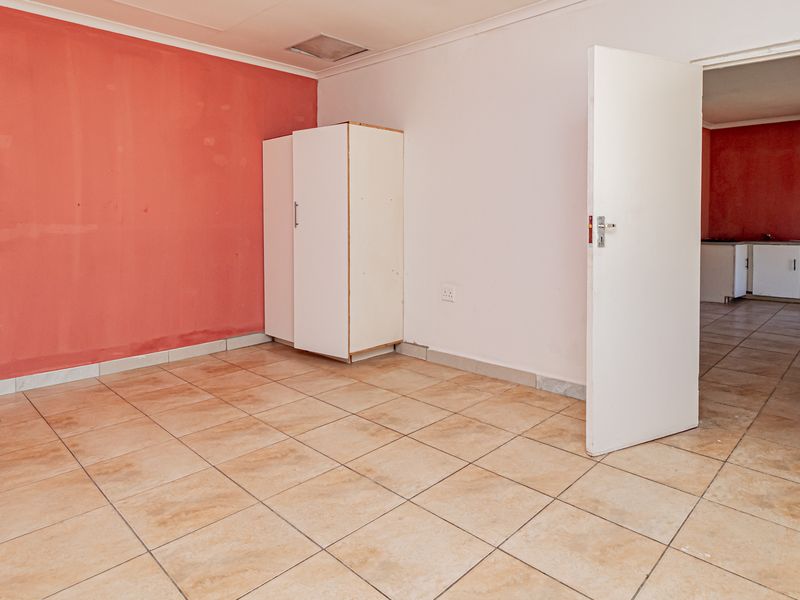 An empty living area featuring ceramic floor tiles and a distinct red accent wall, leading through to an adjacent kitchen space.