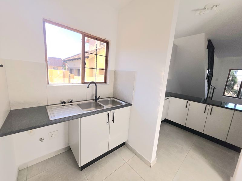 A clean, functional kitchen area featuring white cabinetry, dark laminate countertops, a double stainless steel sink, and neutral tiled flooring.