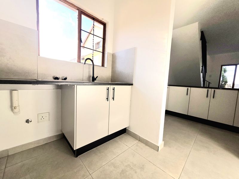 A clean, modern kitchen area featuring white cabinetry, dark countertops, and tiled flooring, with plumbing connections ready for appliances.