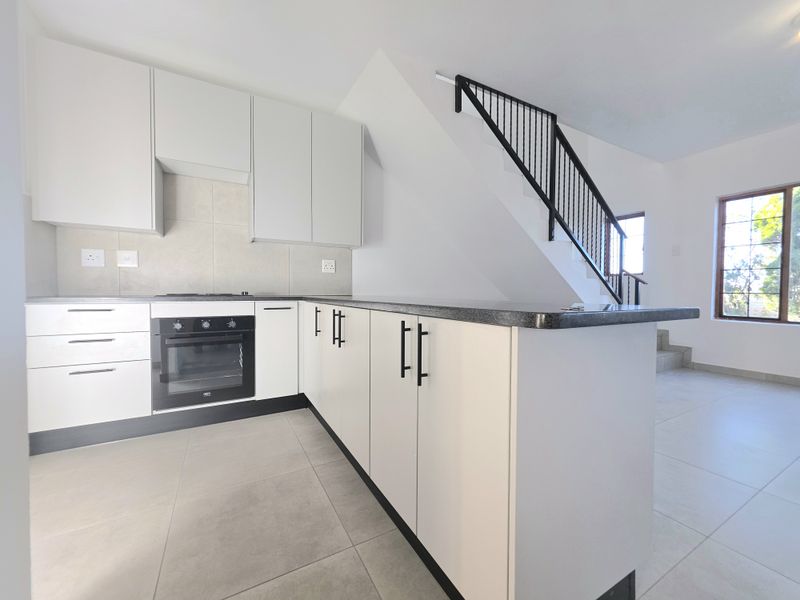 Modern open-plan kitchen featuring sleek white cabinetry, stone countertops, built-in oven, and hob, seamlessly integrated with a staircase area and tiled flooring.