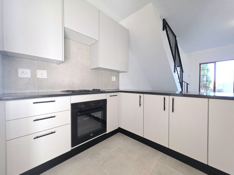 Modern kitchen featuring sleek white cabinetry, dark stone countertops, built-in oven, and hob, situated adjacent to an internal staircase.