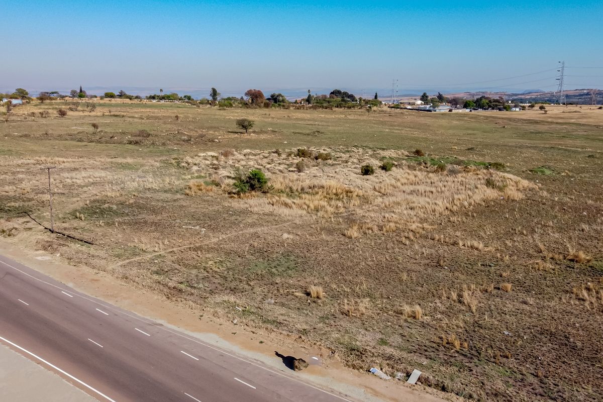 An aerial view of a large, undeveloped vacant residential plot adjacent to a paved road, featuring natural grassland and scattered vegetation.
