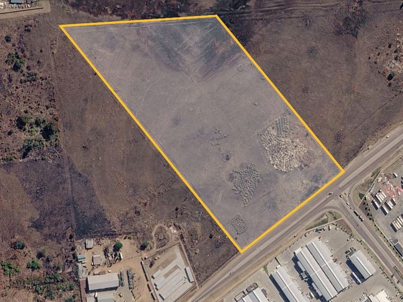 An aerial view of a vacant residential land parcel highlighted with a yellow boundary, situated near commercial infrastructure and main road access.