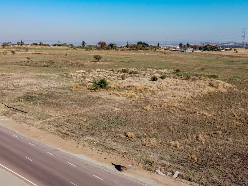 An aerial view of a large, undeveloped vacant residential plot adjacent to a paved road, featuring natural grassland and scattered vegetation.