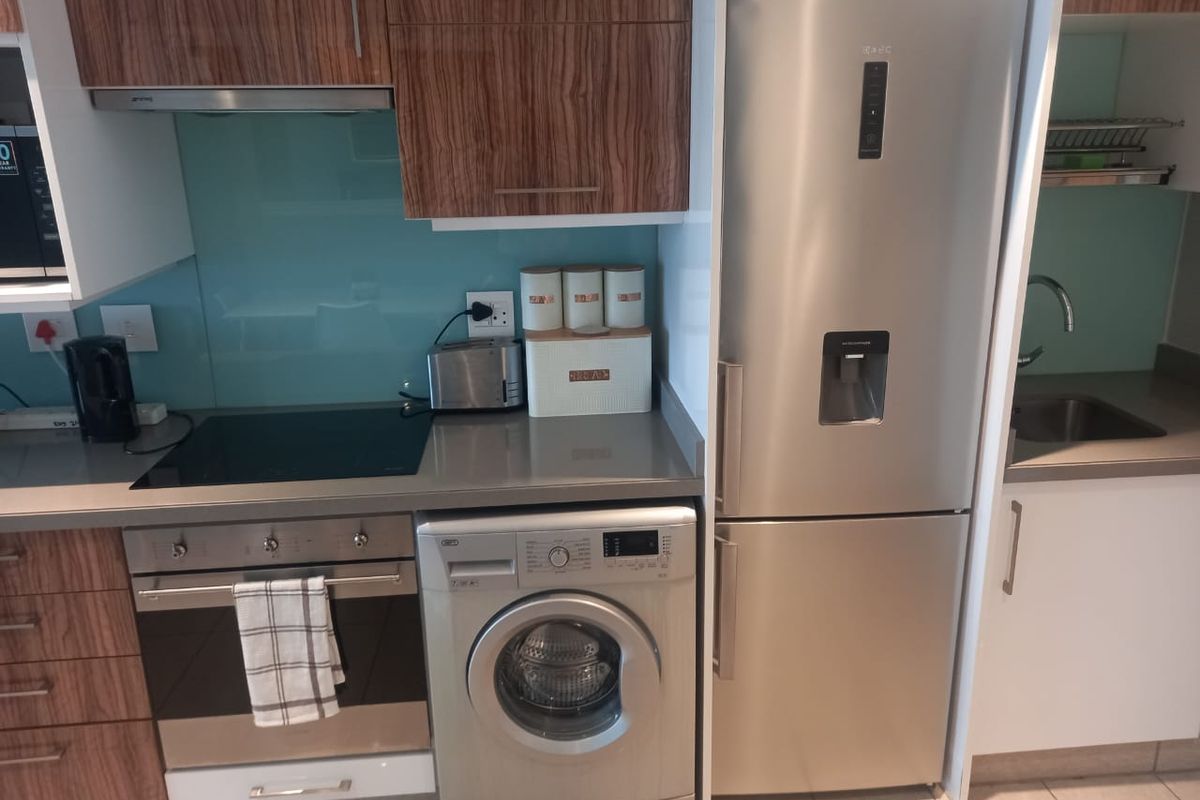 Modern kitchen featuring wood-grain cabinetry, stone countertops, a glass splashback, built-in oven, hob, extractor fan, and integrated laundry appliance space.