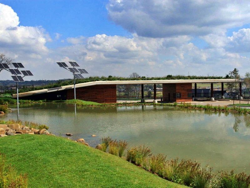 Modern architectural building featuring a green roof, timber cladding, and integrated solar panels, set alongside a landscaped pond and garden area.