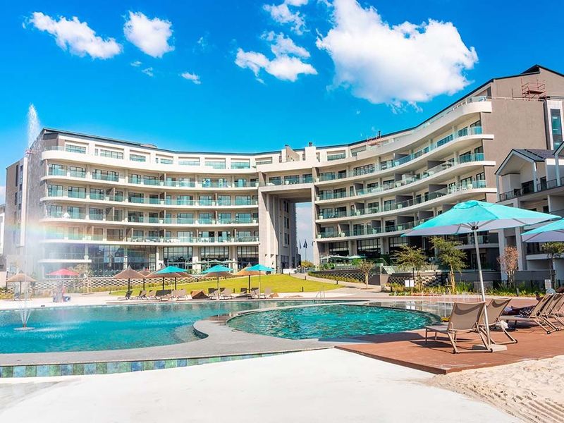 A well-maintained apartment complex featuring a large swimming pool with a water fountain, wooden decking, and landscaped garden areas.