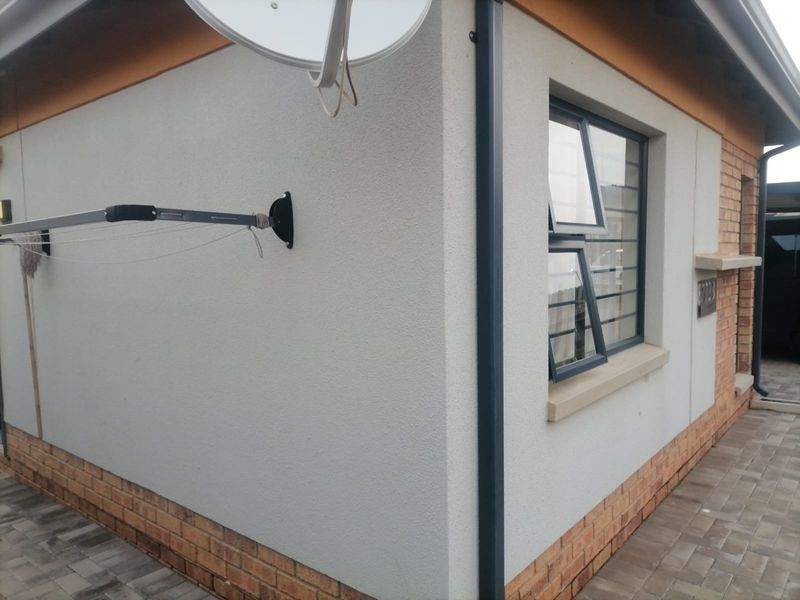 A view of the property exterior featuring a clean plastered facade, brick plinth, modern aluminium window frames, and paved walkway.